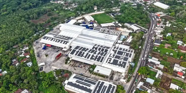 Vista aérea de una gran agencia empresarial con paneles solares instalados en el techo, rodeada de vegetación y una carretera con tráfico.