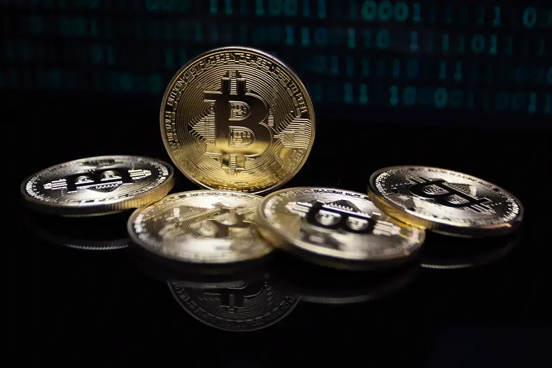 Moneda dorada de Bitcoin erguida rodeada de otras monedas sobre una superficie negra con reflejos y código binario.