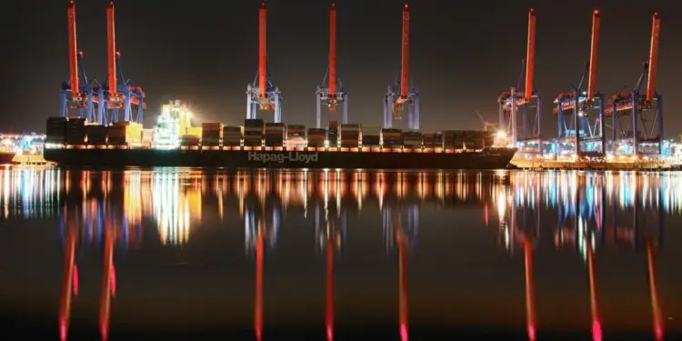 Buque portacontenedores Hapag-Lloyd cargado en un puerto iluminado de noche, con grúas de carga y reflejos vibrantes en el agua.