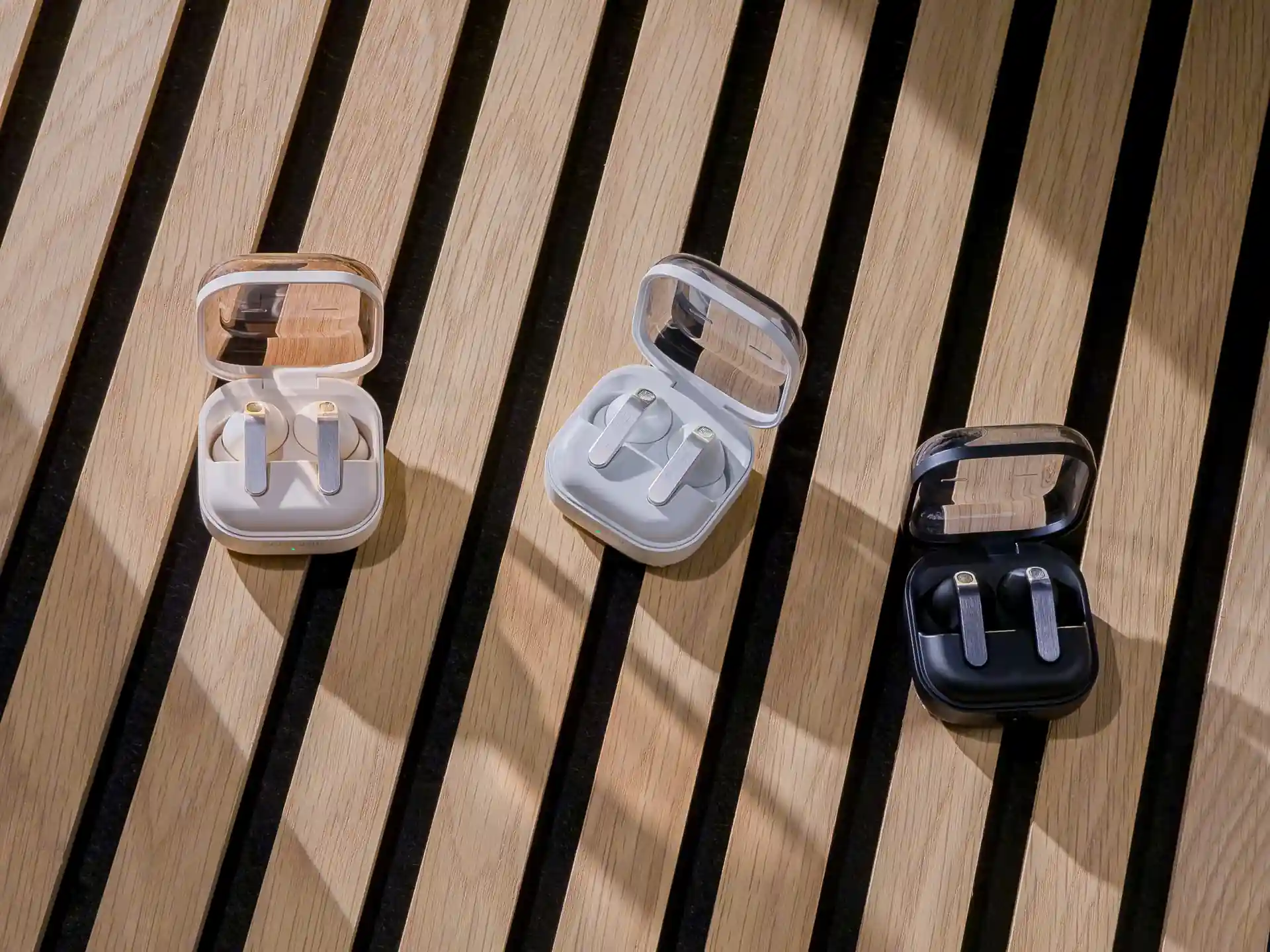 Tres estuches abiertos con auriculares inalámbricos en colores beige, blanco y negro sobre un fondo de listones de madera.