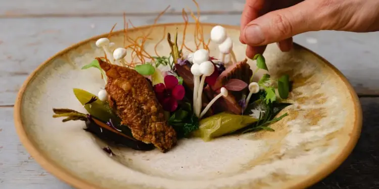 Chef emplatando con cuidado un plato gourmet con hongos blancos, flores comestibles y vegetales en un plato cerámico.
