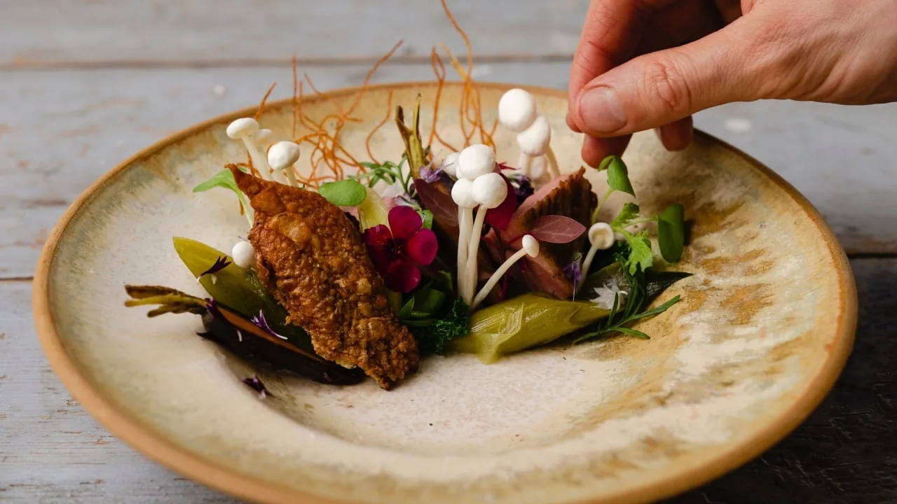 Chef emplatando con cuidado un plato gourmet con hongos blancos, flores comestibles y vegetales en un plato cerámico.