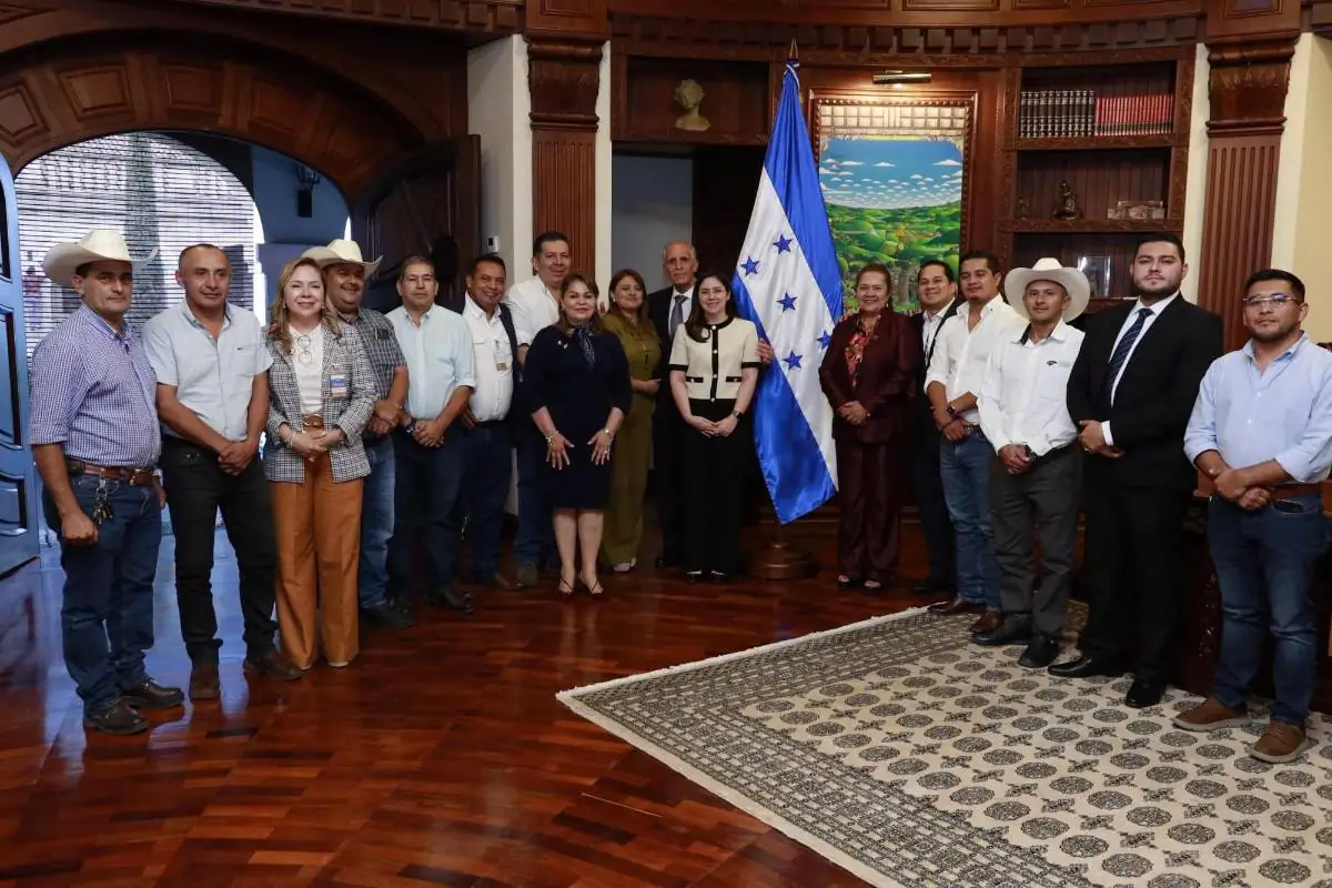 Grupo de hombres y mujeres de negocios y autoridades posando junto a la bandera de Honduras en una sala oficial.