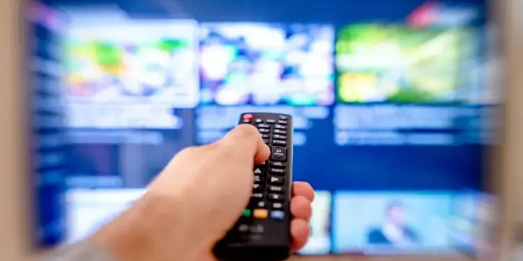 Mano sosteniendo un control remoto frente a una pantalla de televisión encendida con múltiples contenidos.