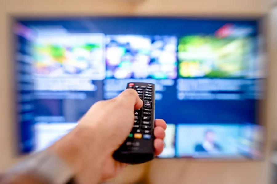 Mano sosteniendo un control remoto frente a una pantalla de televisión encendida con múltiples contenidos.