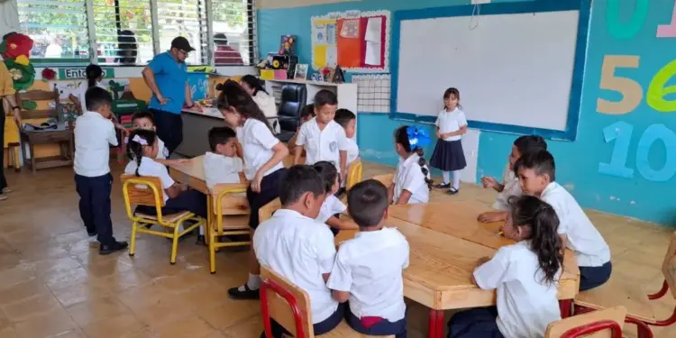 Niños y profesores en un aula escolar realizando actividades educativas sentados en mesas de madera.