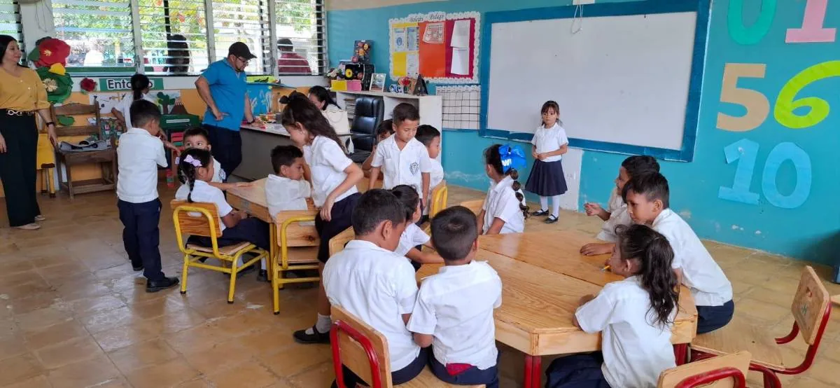Niños y profesores en un aula escolar realizando actividades educativas sentados en mesas de madera.