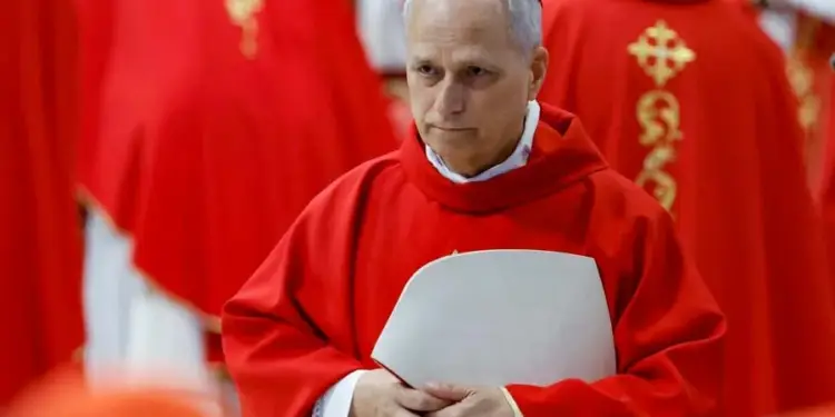 Un hombre con cabello blanco viste túnica roja y un solideo rojo en la cabeza, sosteniendo un documento.