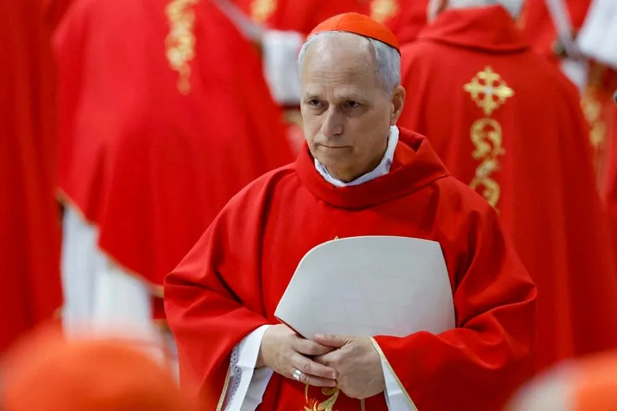Un hombre con cabello blanco viste túnica roja y un solideo rojo en la cabeza, sosteniendo un documento.