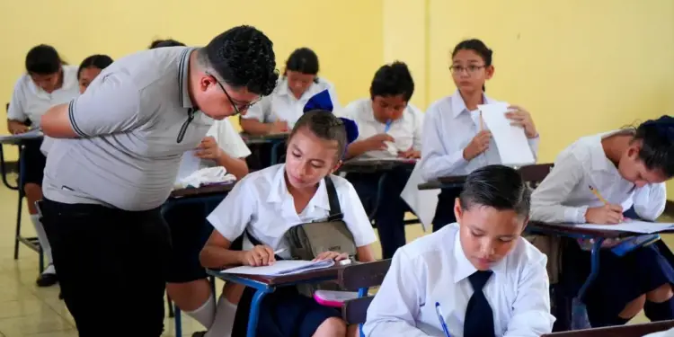Profesor o estudiante de pie revisando el trabajo de sus compañeros en un aula escolar.