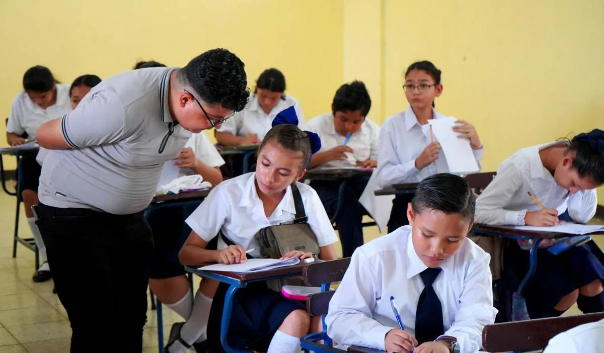 Profesor o estudiante de pie revisando el trabajo de sus compañeros en un aula escolar.