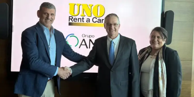 Tres profesionales, dos hombres y una mujer, celebran un acuerdo. Dos hombres se dan la mano frente a una pantalla con los logos de 'UNO Rent a Car' y 'Grupo AM'.
