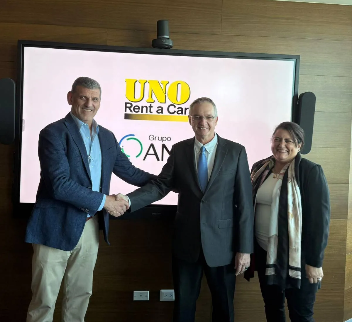 Tres profesionales, dos hombres y una mujer, celebran un acuerdo. Dos hombres se dan la mano frente a una pantalla con los logos de 'UNO Rent a Car' y 'Grupo AM'.