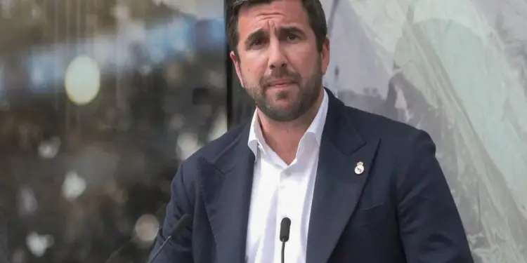 Hombre de negocios en traje oscuro y camisa blanca, con barba, frente a micrófonos en una conferencia. Sujetador con emblema en solapa.