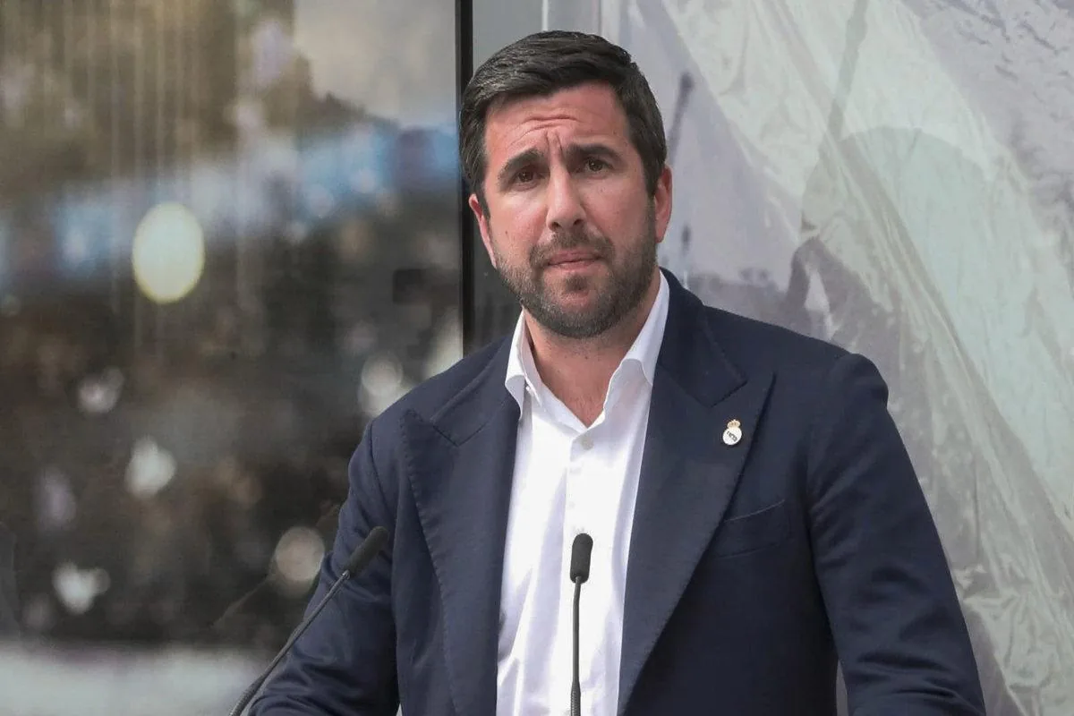 Hombre de negocios en traje oscuro y camisa blanca, con barba, frente a micrófonos en una conferencia. Sujetador con emblema en solapa.