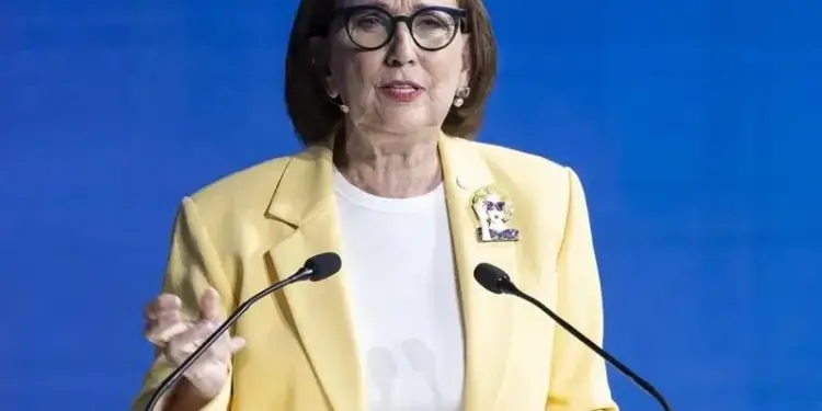 Una mujer de negocios con chaqueta amarilla hablando ante un atril con micrófonos frente a un fondo azul.