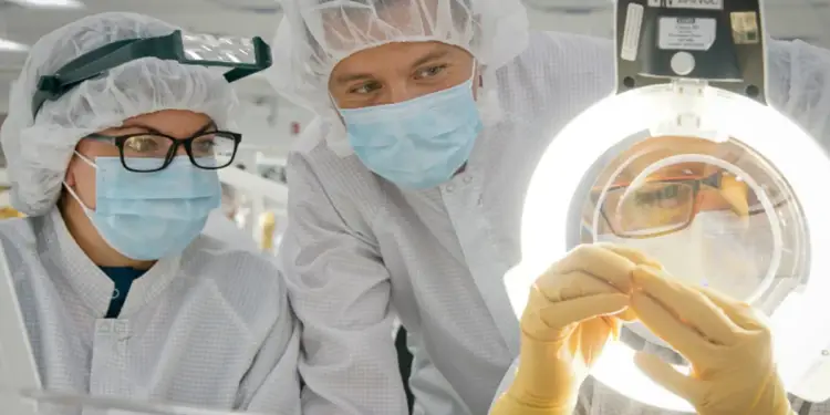 Trabajadores con cofia, mascarilla y guantes inspeccionando componentes bajo una luz de aumento en una planta.