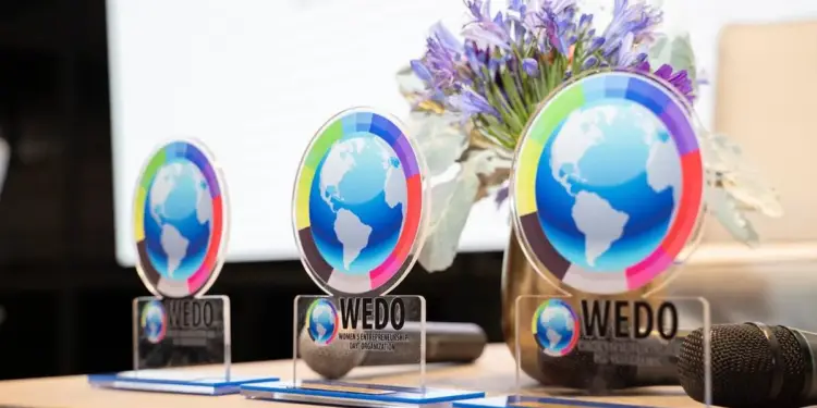 Tres premios de acrílico con un globo terráqueo y segmentos de colores, con el logo WEDO, sobre una mesa de madera junto a flores y un micrófono.