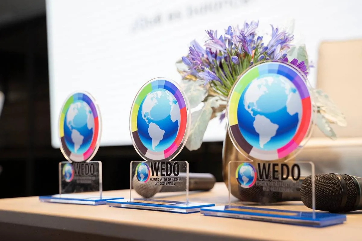 Tres premios de acrílico con un globo terráqueo y segmentos de colores, con el logo WEDO, sobre una mesa de madera junto a flores y un micrófono.