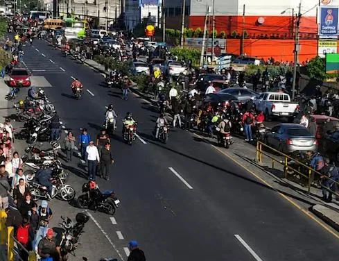 Avenida transitada con numerosos motociclistas, vehículos y peatones en una zona comercial urbana.