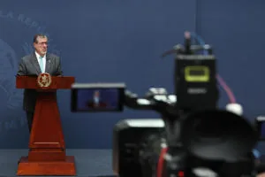 Hombre de traje hablando en un atril de madera frente a cámaras de televisión en una conferencia