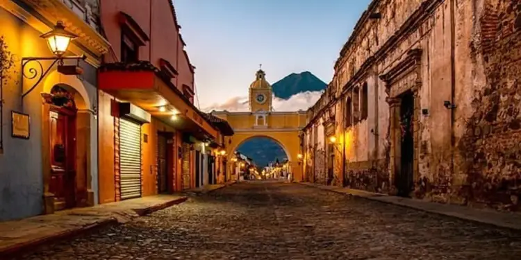 Calle colonial adoquinada flanqueada por edificios históricos coloridos y un arco amarillo con reloj al fondo durante el atardecer