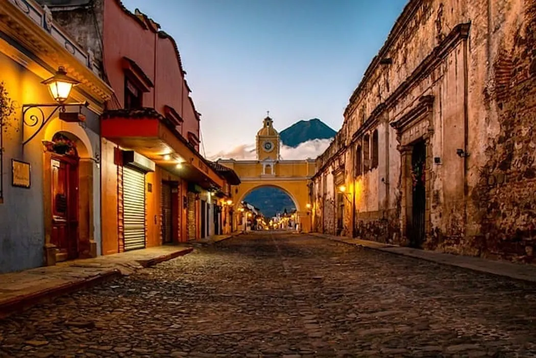Calle colonial adoquinada flanqueada por edificios históricos coloridos y un arco amarillo con reloj al fondo durante el atardecer