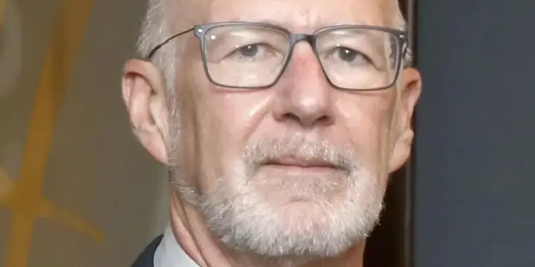 Hombre mayor calvo con gafas, barba cana, traje oscuro y corbata mirando a la cámara.