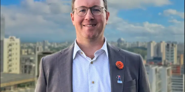 Ejecutivo con gafas y traje gris sonriendo con pin de Hilton, con fondo de edificios urbanos y cielo azul.