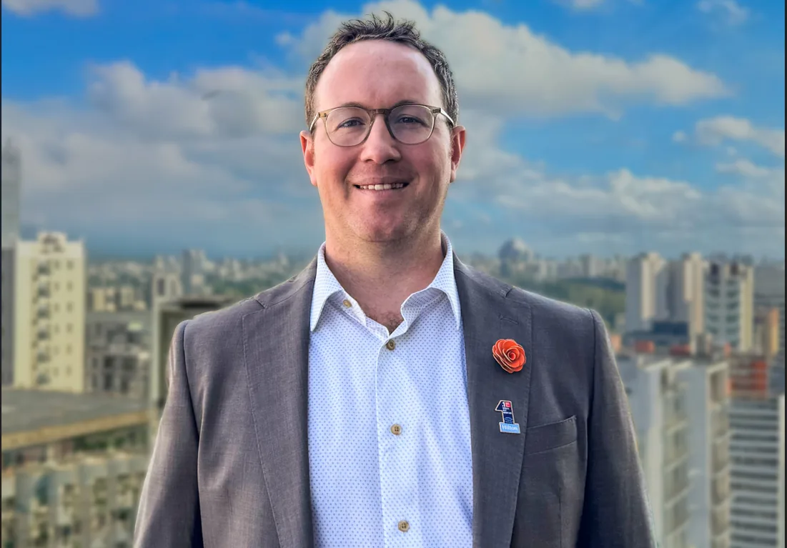 Ejecutivo con gafas y traje gris sonriendo con pin de Hilton, con fondo de edificios urbanos y cielo azul.