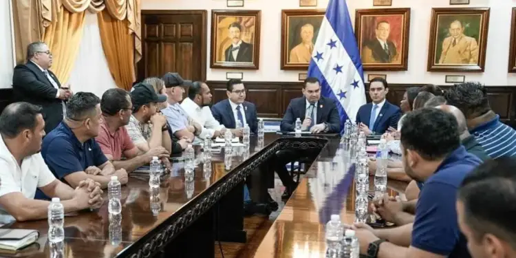 Hombres en una reunión formal alrededor de una mesa de madera pulida, con la bandera de Honduras y retratos en la pared. Uno habla al micrófono.