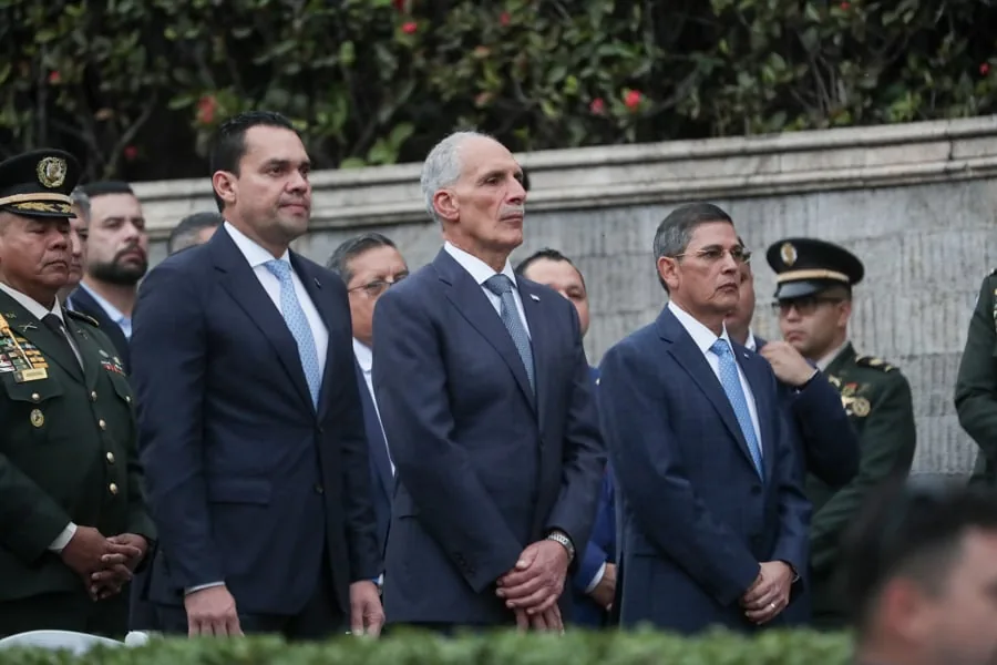 Grupo de hombres con traje formal y oficiales con uniforme militar asistiendo a un evento oficial y protocolario.