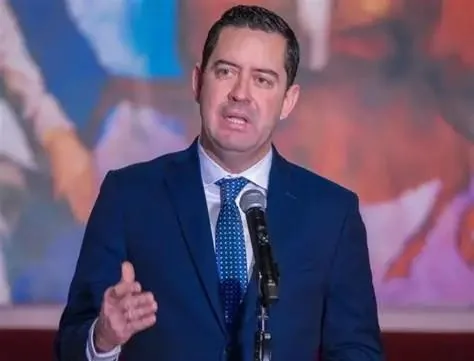 Hombre profesional en traje azul oscuro y corbata, hablando en un micrófono durante un evento. Gesticula con la mano derecha, con un fondo abstracto y colorido.