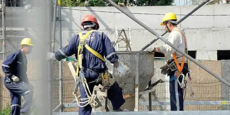 Tres obreros con cascos y arneses trabajando en una obra de construcción junto a un contenedor de mezcla.