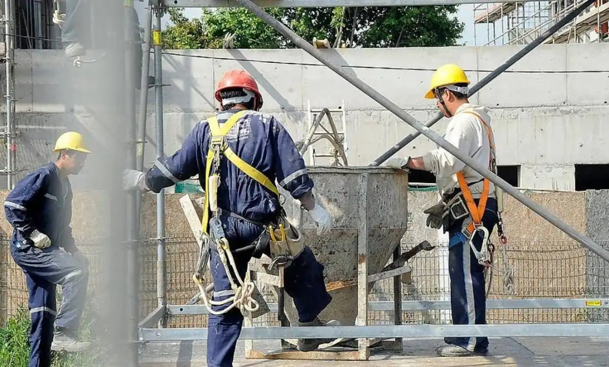 Tres obreros con cascos y arneses trabajando en una obra de construcción junto a un contenedor de mezcla.