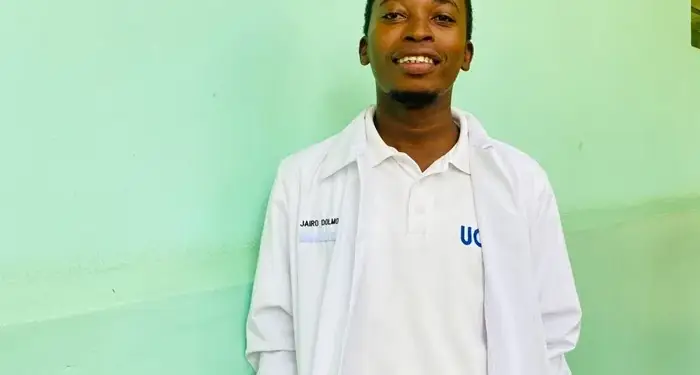 Joven sonriente de pie junto a una pared verde, vistiendo una bata blanca de laboratorio sobre una camisa blanca y pantalón oscuro.
