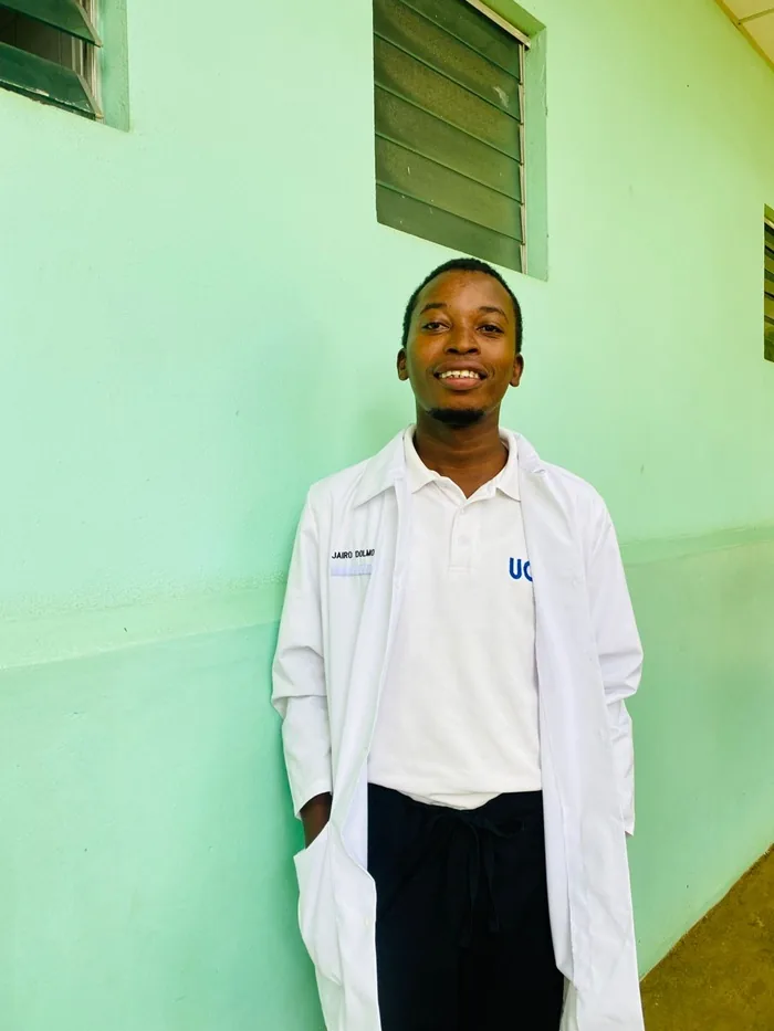 Joven sonriente de pie junto a una pared verde, vistiendo una bata blanca de laboratorio sobre una camisa blanca y pantalón oscuro.