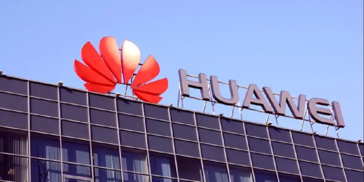 Primer plano del letrero de Huawei en el tejado de un edificio moderno con fachada oscura y ventanas, bajo un cielo azul claro. El logo rojo y el nombre 'HUAWEI' son prominentes.