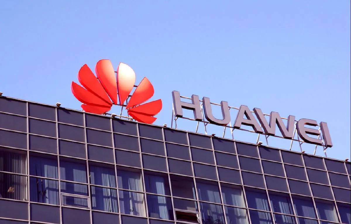 Primer plano del letrero de Huawei en el tejado de un edificio moderno con fachada oscura y ventanas, bajo un cielo azul claro. El logo rojo y el nombre 'HUAWEI' son prominentes.
