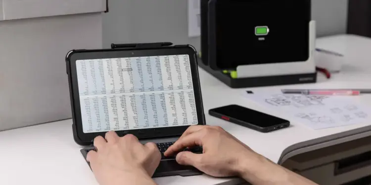 Persona escribiendo en un teclado conectado a una tablet que muestra hojas de cálculo financieras en una oficina