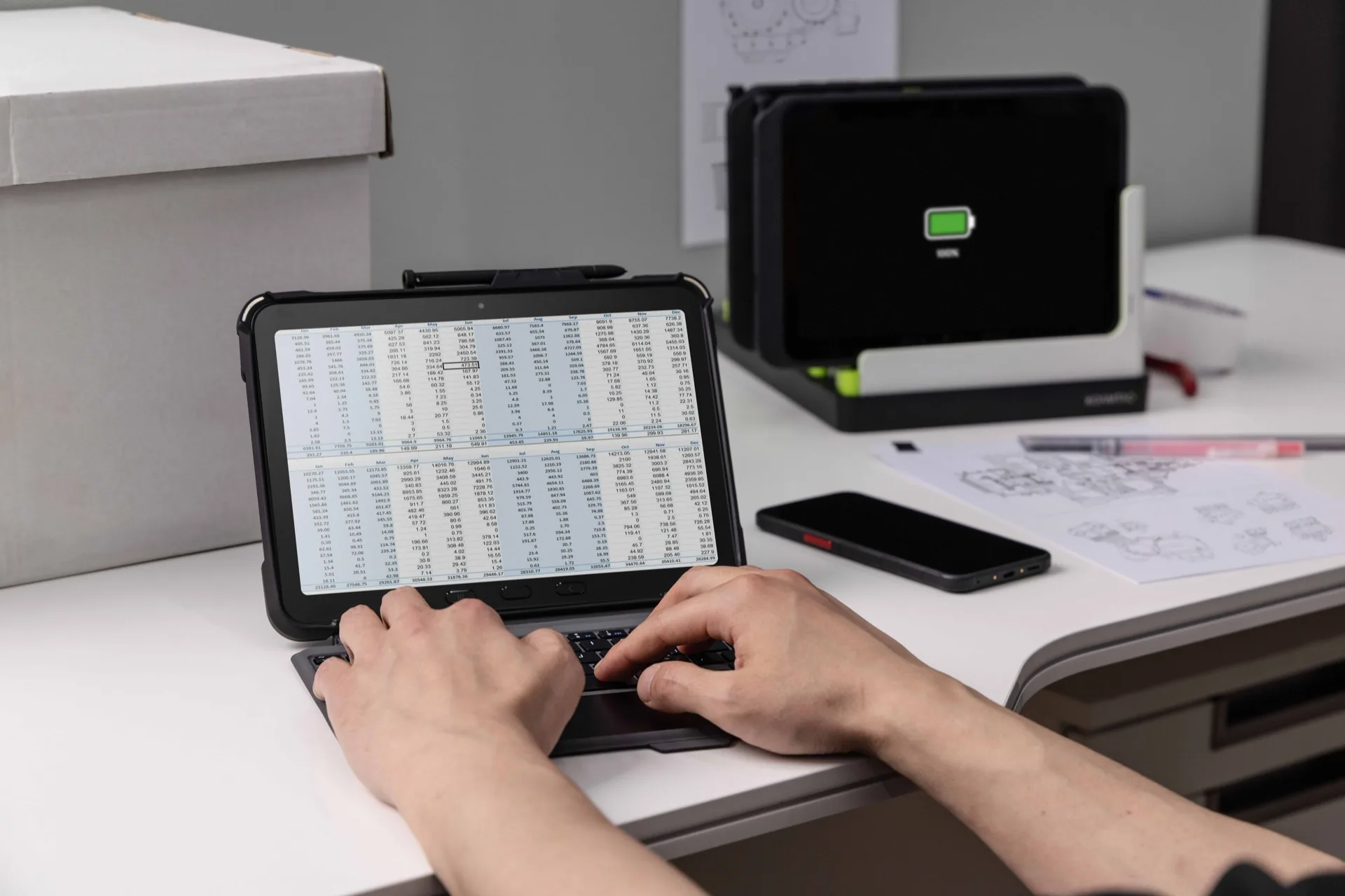 Persona escribiendo en un teclado conectado a una tablet que muestra hojas de cálculo financieras en una oficina