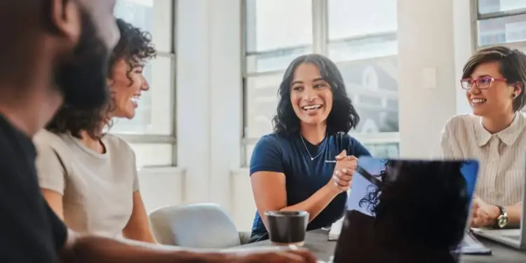 Cuatro profesionales sonrientes en una reunión de negocios, colaborando en una oficina moderna con laptops y tazas de café.