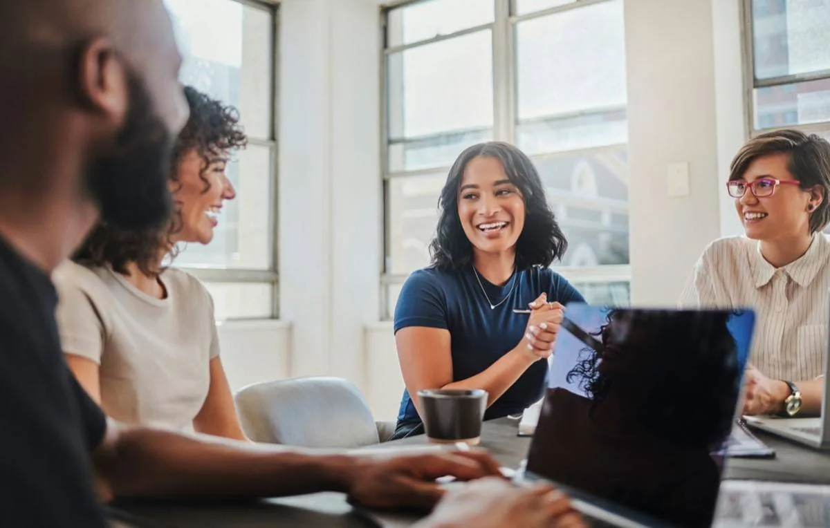 Cuatro profesionales sonrientes en una reunión de negocios, colaborando en una oficina moderna con laptops y tazas de café.