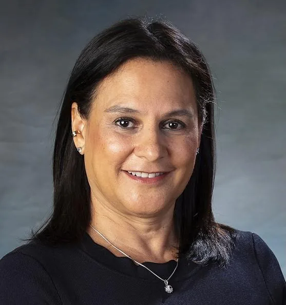Retrato de media distancia de una mujer sonriente con cabello oscuro y blusa negra sobre fondo gris.