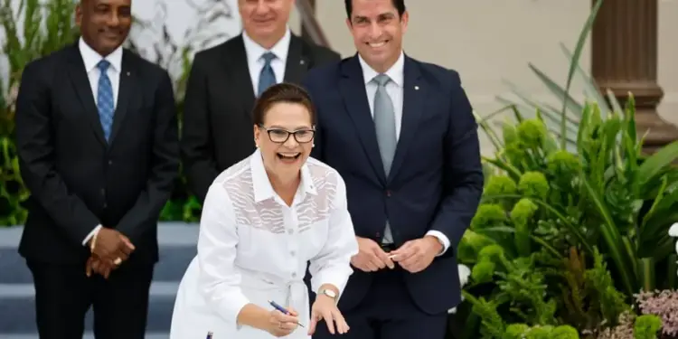 Mujer sonriente firmando un documento en una mesa de cristal, con tres hombres en traje detrás. Escena de acuerdo o colaboración profesional.