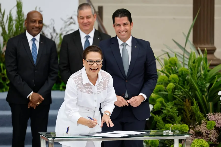 Mujer sonriente firmando un documento en una mesa de cristal, con tres hombres en traje detrás. Escena de acuerdo o colaboración profesional.