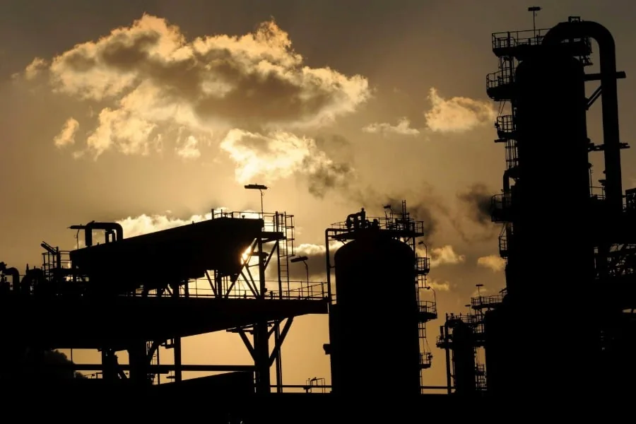 Silueta de una refinería o planta industrial con grandes torres y estructuras metálicas contra un cielo al atardecer.