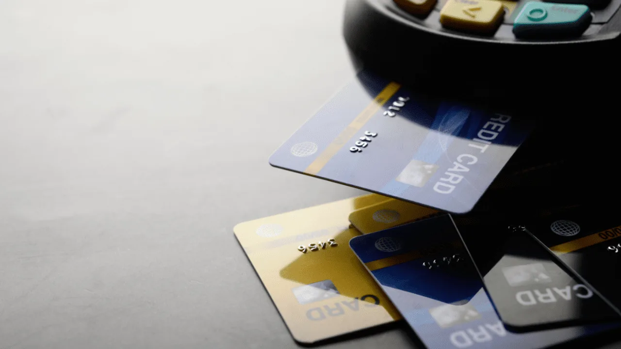 Varias tarjetas de crédito (azul, dorado, negro) con una insertada en un terminal de punto de venta (POS). Teclas visibles.