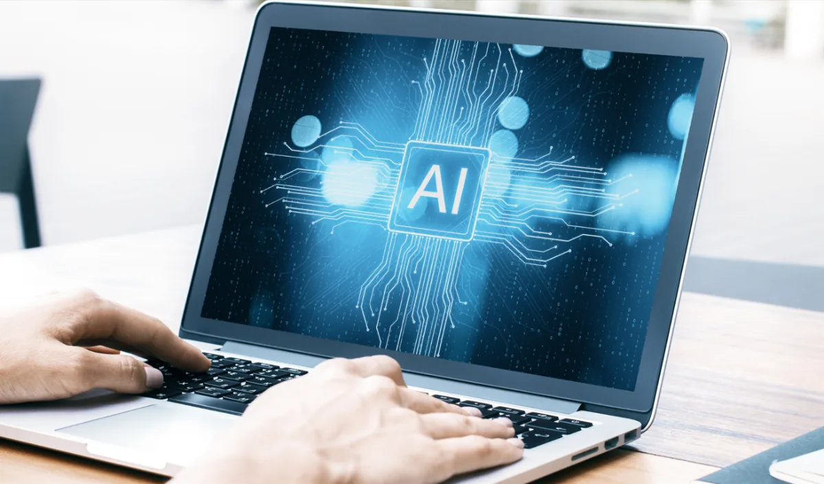 Primer plano de manos escribiendo en un portátil con una pantalla que muestra el logo de IA rodeado de circuitos digitales y luces bokeh azules.