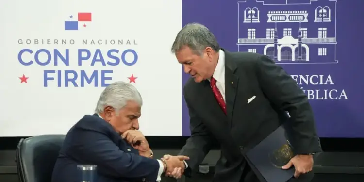 Dos hombres en traje estrechando las manos en un evento oficial con paneles institucionales al fondo.